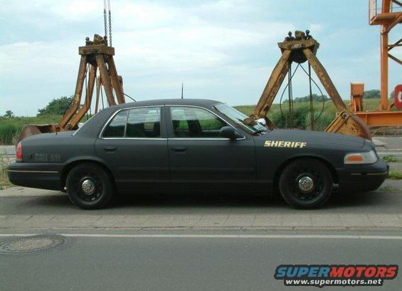 Side view of a black Ford Crown Victoria with "SHERIFF" on the door and "CALL 911" on the rear quarter panel. It's parked in front of industrial equipment