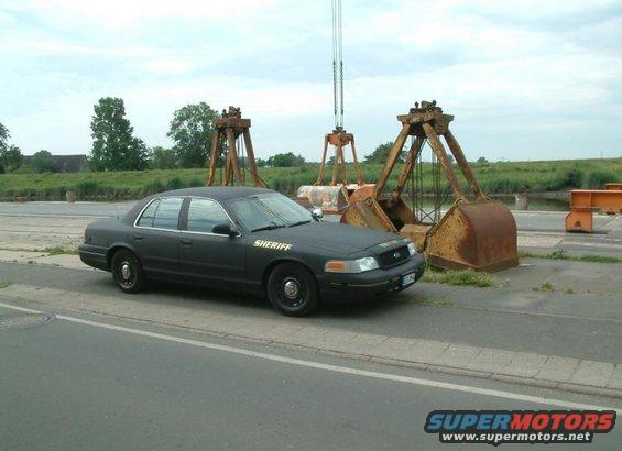 Black Ford Crown Victoria police car with "SHERIFF" on the hood, parked near industrial equipment