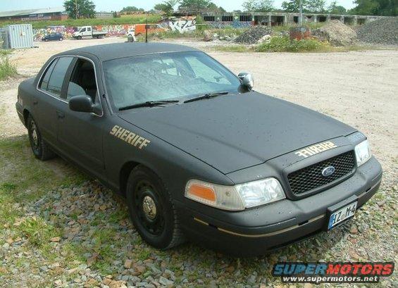Black Ford Crown Victoria with 'SHERIFF' on the door and hood, parked on gravel