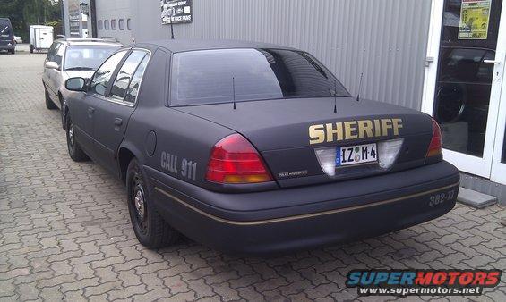 Black matte sheriff car with gold 'SHERIFF' text and 'CALL 911' on the side, parked on a brick driveway