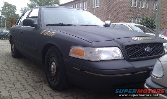 Matte black 2003 Ford Crown Victoria police car with 'SHERIFF' text on the hood and door. Another car is partially visible in the foreground