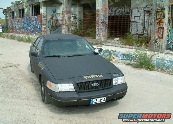 2003 Ford Crown Victoria police car with "SHERIFF" text on the fender and "382-17" on the hood, parked in front of a graffiti-covered building