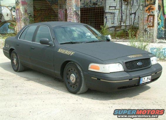 Black 2003 Ford Crown Victoria sheriff car with gold lettering on the fender and door, parked on dirt in front of a graffiti-covered building