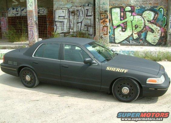 Black 2003 Ford Crown Victoria with 'SHERIFF' on the hood and 'CALL 911' on the rear fender, parked in front of graffiti-covered building