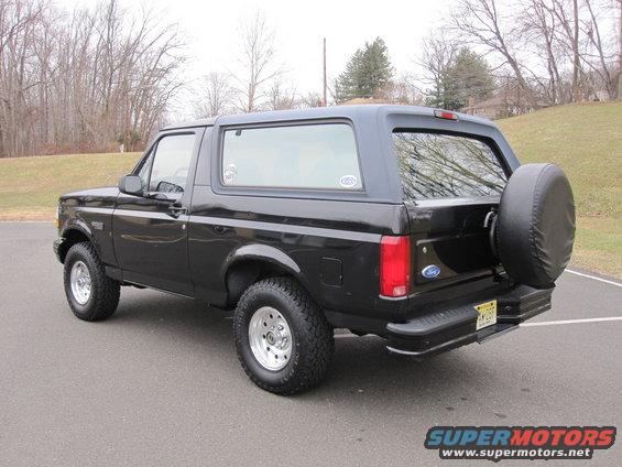 Black Ford Bronco parked on asphalt with trees and a grassy hill in the background. Spare tire mounted on the rear