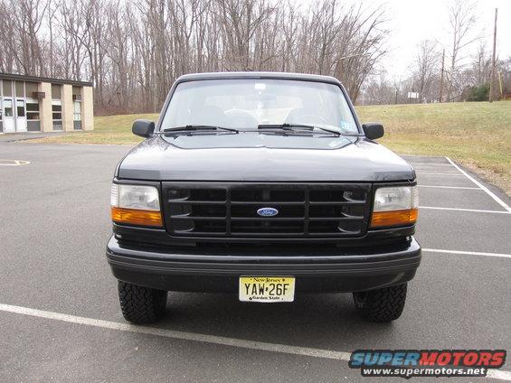 Black Ford Bronco front view, parked in a lot with a building and trees in the background