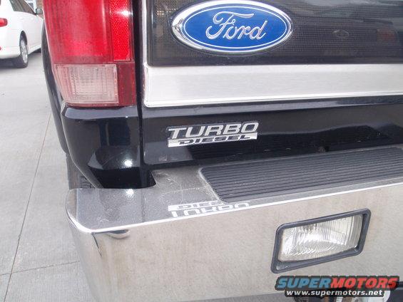 Black Ford truck rear bumper with chrome accents and a "Turbo Diesel" badge. A white car is visible to the left