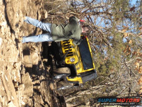 Yellow Jeep Wrangler tilted on a rocky incline, driver's side visible, with a person in jeans and a green jacket standing nearby