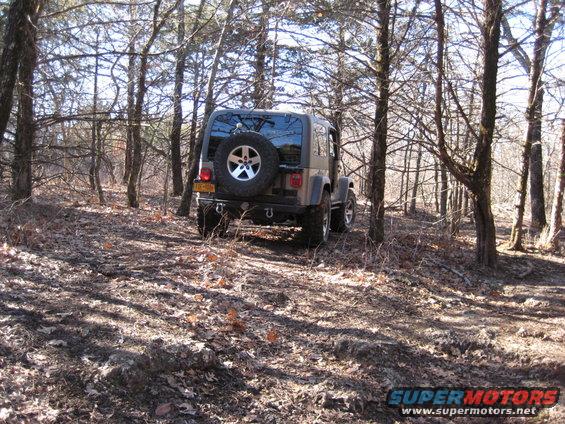 Brown Jeep Wrangler with spare tire on rear, driving on a dirt path through trees