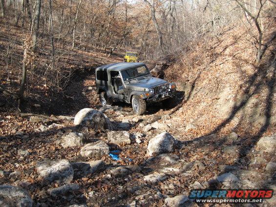 Gray Jeep Wrangler navigating a rocky, wooded trail with a yellow Jeep in the background