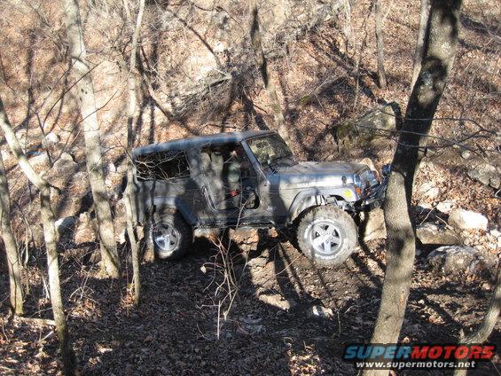 Gray Jeep Wrangler on rocky, wooded terrain, driver's door open
