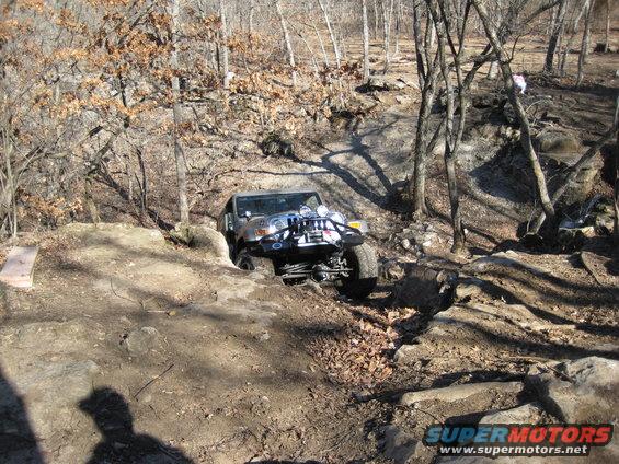Jeep Wrangler navigating a rocky, wooded trail. The vehicle is angled, with its front wheels turned