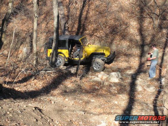 Yellow 2005 Jeep Wrangler navigating a rocky, wooded trail with a person standing nearby