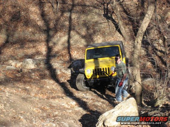 Yellow Jeep Wrangler driving on a rocky, wooded trail with a person standing nearby