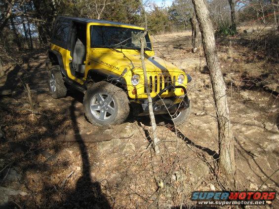 Yellow Jeep Wrangler Rubicon driving on a rocky, wooded trail. The front passenger door is open