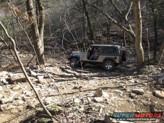 Jeep Wrangler driving on a rocky trail in a wooded area. Driver is visible