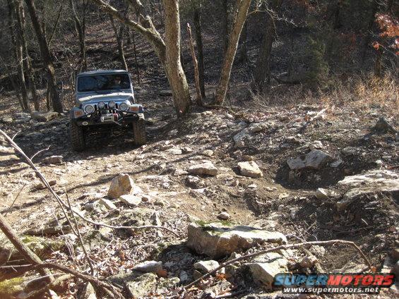 Silver Jeep Wrangler driving up a rocky hill in a wooded area. It has a winch on the front bumper