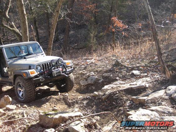 Jeep Wrangler Rubicon driving over rocks on a wooded trail. It has a winch and extra lights on the front