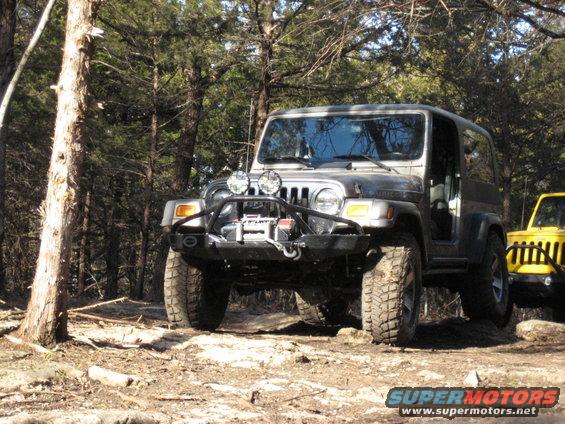 Gray Jeep Wrangler with winch and auxiliary lights on a rocky trail, with a yellow Jeep visible behind it