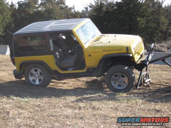Yellow Jeep Wrangler with hardtop, driver's door open, parked on dry grass. Rubicon badge visible on hood