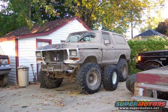 Mud-covered 1978 Ford Bronco with large tires and dual rear wheels parked next to a garage. Another black truck is visible to the right