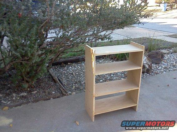 Unfinished plywood shelving unit on a concrete sidewalk next to a bush