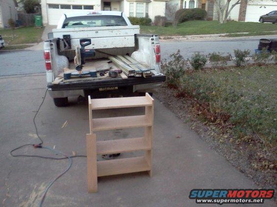 Pickup truck bed loaded with lumber, a miter saw, and tools. A wooden shelf unit is in the foreground