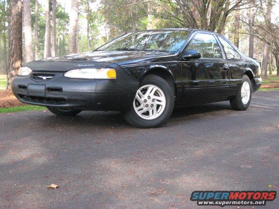 Black Ford Thunderbird parked on asphalt with trees in background. Has a car bra on the front