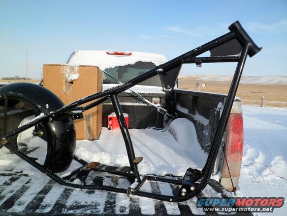 Black motorcycle frame in snow-covered truck bed with a tire and boxes