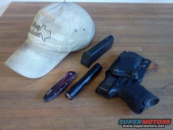 Hat with Texas outline, folding knife, tactical flashlight, handgun in holster, and spare magazine on wooden surface