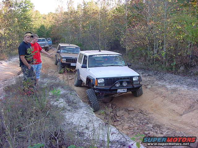 White Jeep Cherokee XJ with a black bumper drives on a dirt trail, followed by other off-road vehicles