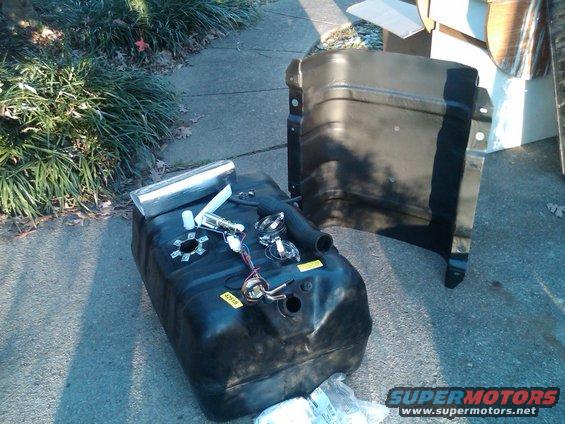 Black plastic fuel tank with fuel pump assembly and metal filler neck, next to a black metal fender liner
