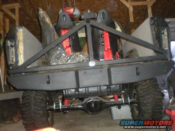Rear view of an off-road vehicle with a custom heavy-duty bumper, roll cage, and racing seats. Leaf springs and differential are visible