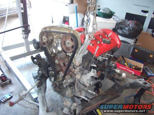 Engine assembly suspended by a chain hoist, with a red valve cover and timing belt visible