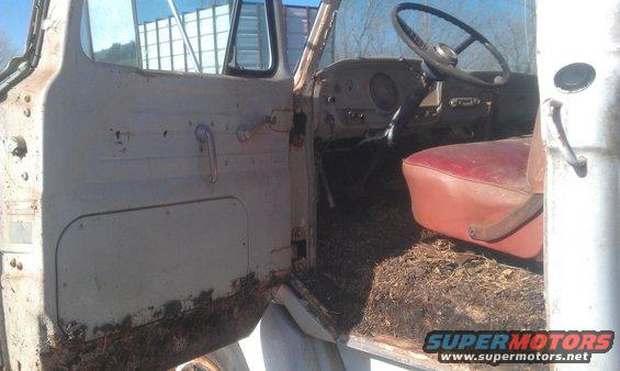 view into an old truck cab, showing the driver's seat, steering wheel, and dashboard. The floor is covered in debris