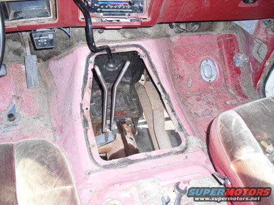 Interior floor of a vehicle with exposed transmission tunnel and worn seats. A "Supermotors.net" watermark is visible