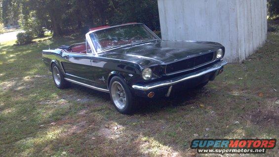 Black 1965 Ford Mustang convertible with red interior, parked on grass near a concrete wall