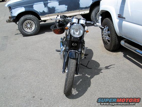 Front view of a motorcycle parked between two trucks. One truck is dark blue with primer patches, the other is white