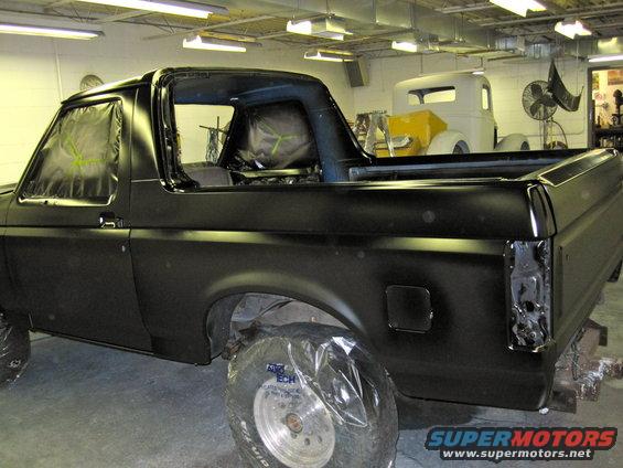 Black Ford Bronco in a shop, windows taped off, ready for paint. Another truck is in the background