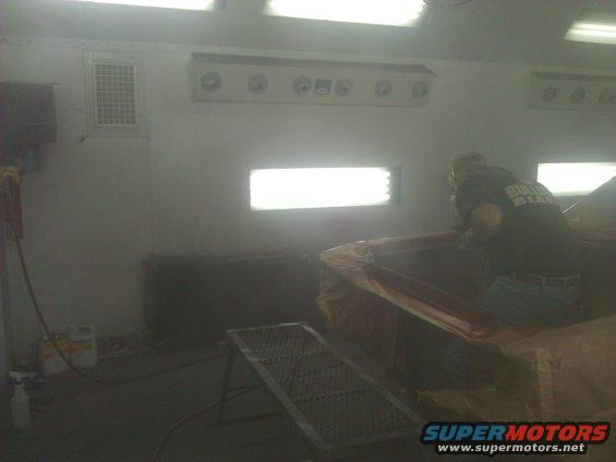 Car interior covered in plastic sheeting inside a brightly lit paint booth with a person working on it