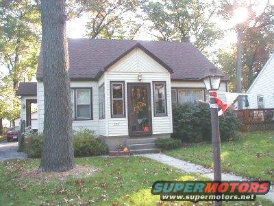 House with a large tree in the foreground and a red car parked in the driveway