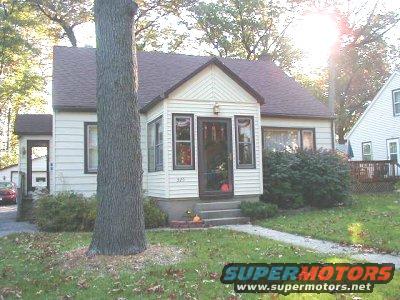House with a large tree in front, driveway with a red car visible
