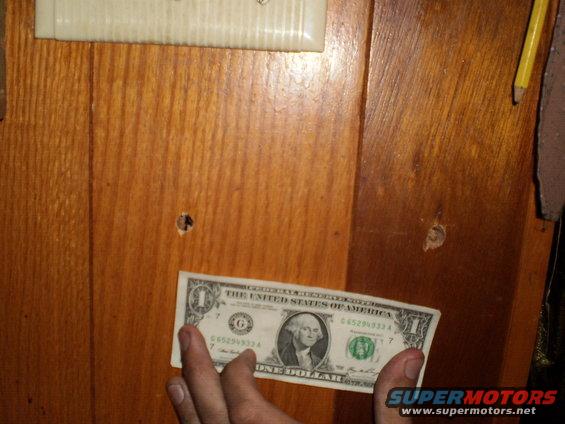 Dollar bill held in front of wood paneling with a pencil on the right