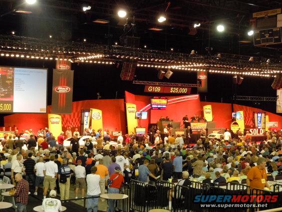 Big crowd watching an auction with a car on stage. A sign shows G109 and $25,000