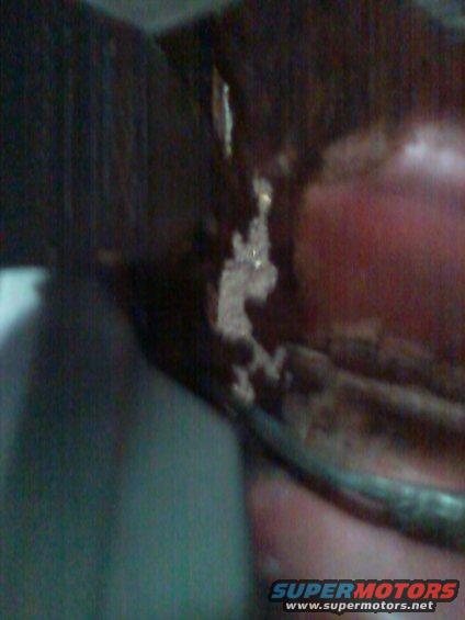 Close-up of a dark-colored vehicle part with some white residue and a red surface behind it. Watermark reads SUPERMOTORS