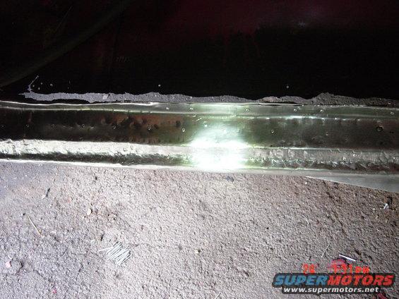 Undercarriage of a vehicle, showing rust and damage along the rocker panel. A bright light illuminates the area