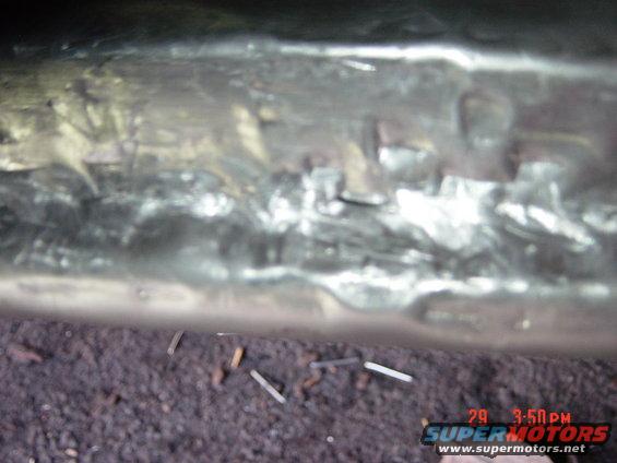 Close-up of a rusty, possibly repaired metal frame under a vehicle. Some debris is on the ground below