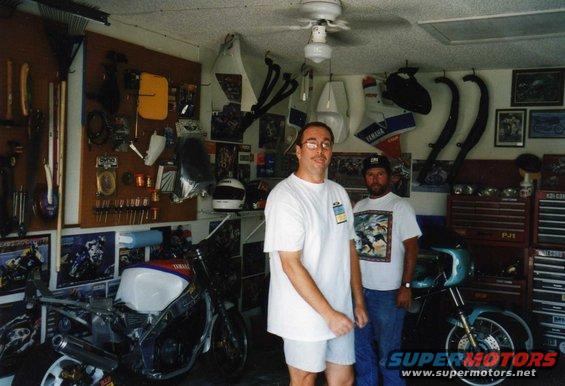Two men stand in a garage with motorcycles and parts visible