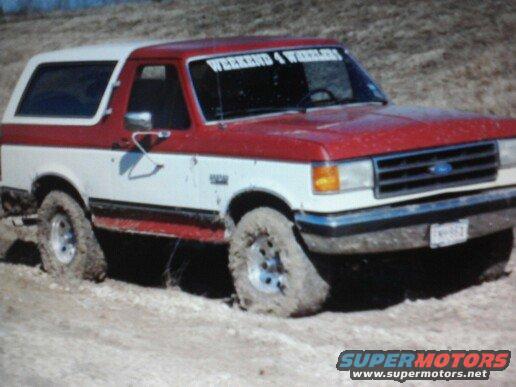 Red and white Ford Bronco stuck in mud, front 3/4 view. "WEEKEND 4 WHEELERS" sticker on windshield