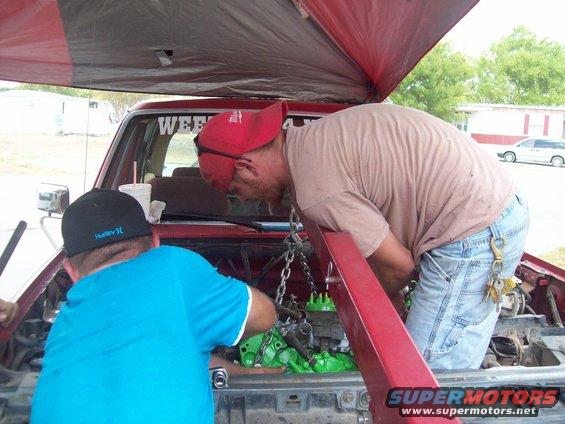 Red vehicle engine bay with two men working on the engine, which is painted green